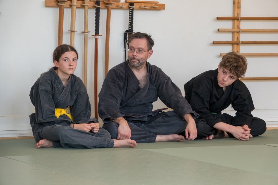 Un instructeur du dojo assis entre deux jeunes pratiquants en keikogi, devant le râtelier de sabres en bois