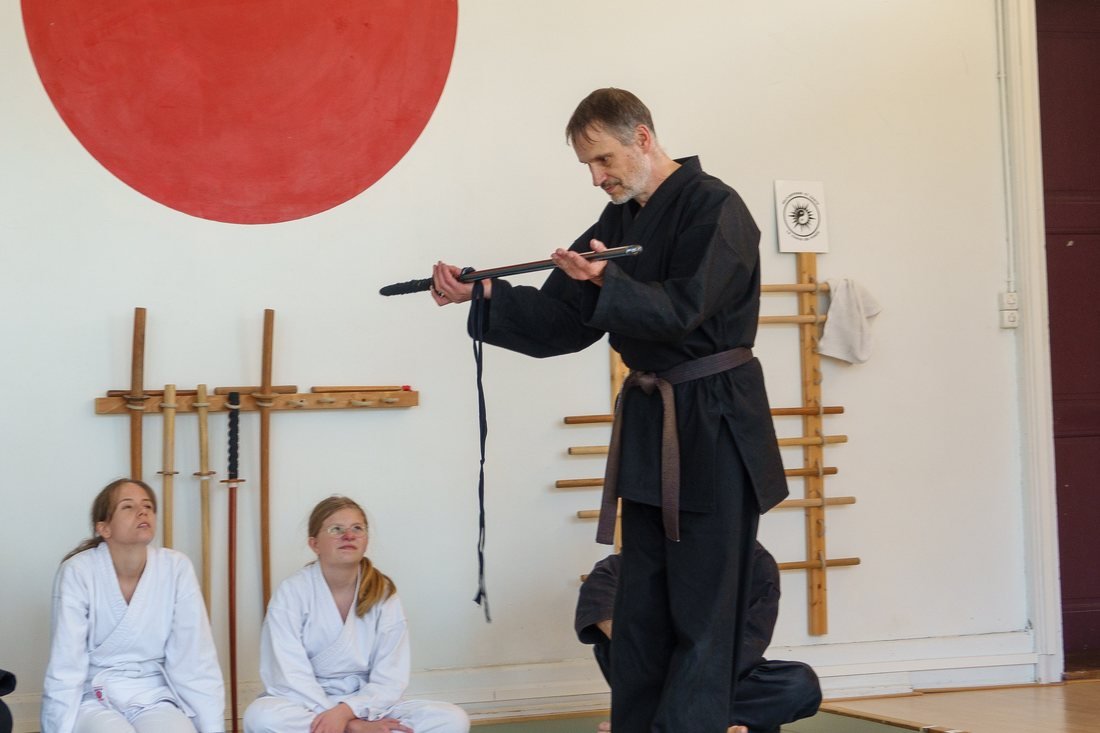 Démonstration au sabre devant le hinomaru rouge du dojo, deux jeunes pratiquants en kimono blanc observant assis